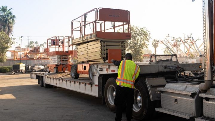 A worker in a safety vest uses telematics to guide orange scissor lifts onto a flatbed truck at an outdoor industrial site.