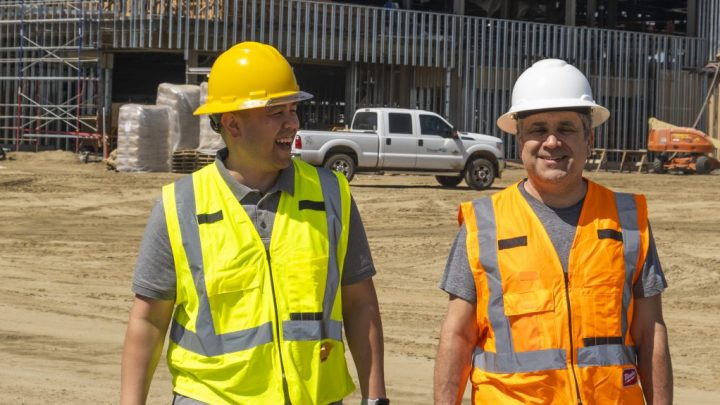 Two construction workers in safety gear walking at a construction site, with telematics-equipped equipment and truck in the background.