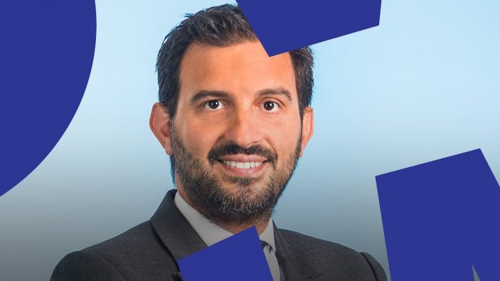 A man with dark hair and a beard, wearing a suit and smiling, stands before a blue background evoking connected jobsite themes.