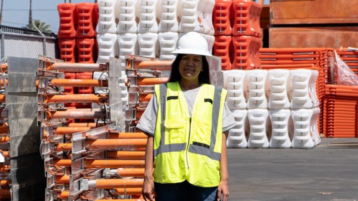 A woman in a white hard hat and yellow vest stands by orange and white construction barriers, overseeing telematics operations.