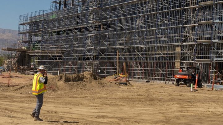 A construction worker uses telematics on a phone at a site with scaffolding, materials, and equipment in view.