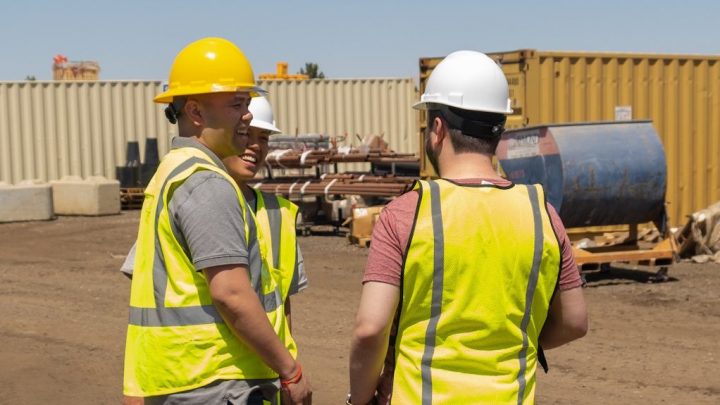 Three construction workers in safety vests and hard hats talk and smile on a dirt worksite with telematics equipment nearby.