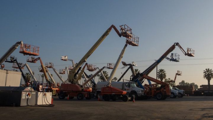 Cherry picker lifts and construction vehicles from a fleet management service parked at sunrise, with palm trees and materials behind.