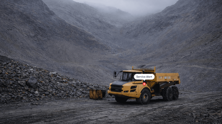 A yellow dump truck with aftermarket parts is parked on a rocky quarry road under a Service Alert, showing construction IoT in action.