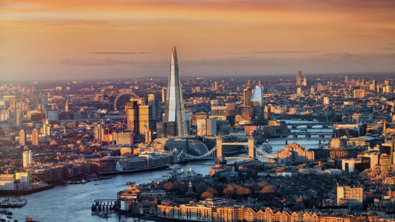 Aerial view of London at sunset, showing the Shard, Tower Bridge, and the River Thames winding through the city under a vibrant orange sky—a glimpse of a skyline set to transform with IPAF elevation UK 2026 initiatives in connected jobsite development.