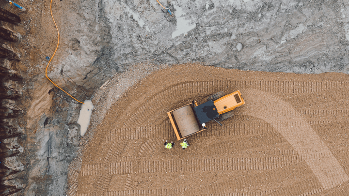 Aerial view of a connected jobsite with a yellow bulldozer moving gravel, two workers in green vests, and tire tracks in the dirt.