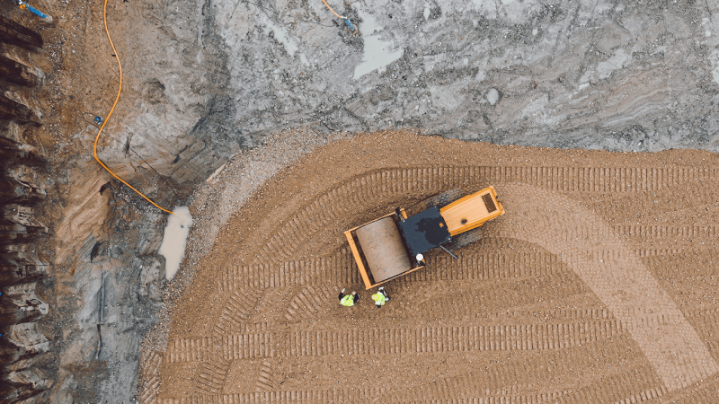 Aerial view of a connected jobsite with a yellow bulldozer moving gravel, two workers in green vests, and tire tracks in the dirt.
