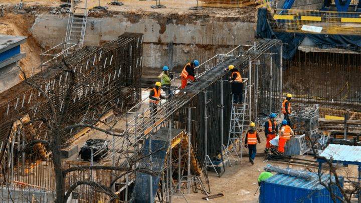 Construction workers in safety gear assemble steel frameworks as construction IoT monitors equipment and materials on site.