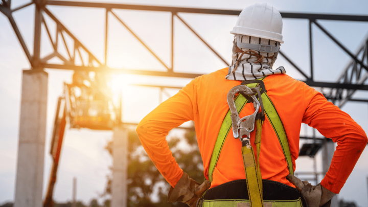 A construction worker in safety gear watches a steel structure rise at sunset, highlighting contractor safety and the connected jobsite.
