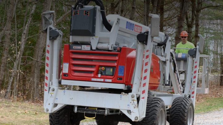 A worker in a hard hat uses a red and white Dino 180XT lift from Sunbelt Rentals on a gravel path, showing construction equipment tracking.