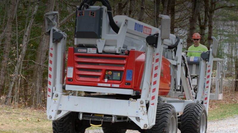 A worker in a hard hat uses a red and white Dino 180XT lift from Sunbelt Rentals on a gravel path, showing construction equipment tracking.