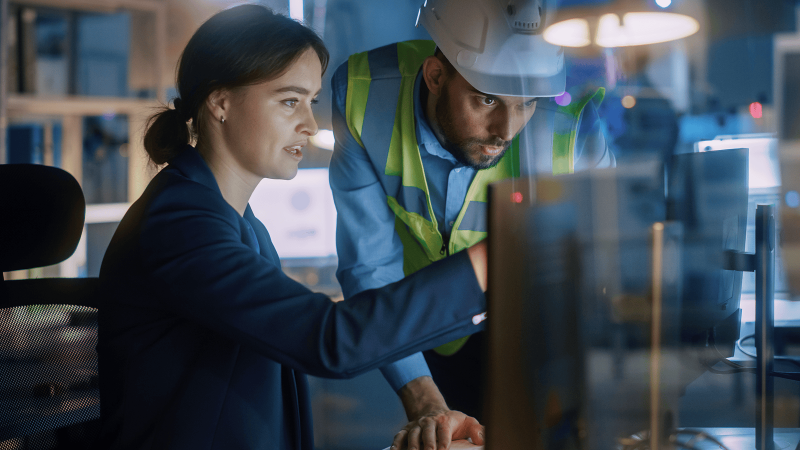 A woman in business attire and a man in a safety vest and hard hat review information together on a computer in an industrial office, collaborating on a construction IoT platform.