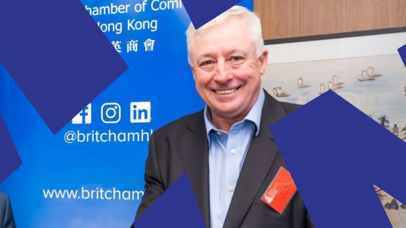 A smiling man in a suit stands before a blue tech-themed banner; dark shapes and a painting with ships hint at fleet management.