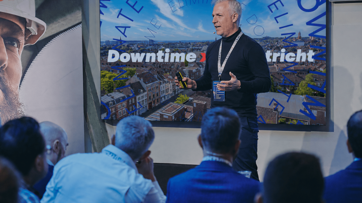 A man in dark clothing speaks on stage at the Downtime Summit; a screen shows "Downtime Maastricht" and an aerial city view, related to fleet management.