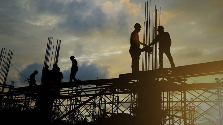 Silhouetted construction workers on scaffolding at sunset, steel rods visible, illustrating downtime's impact on a connected jobsite.