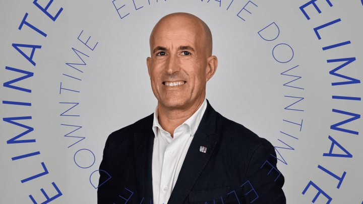A smiling bald man in a dark suit poses against a gray backdrop with blue text, "ELIMINATE DOWNTIME," highlighting telematics.