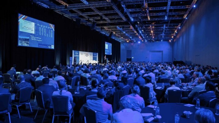 Conference room with attendees at round tables, stage speakers, blue lighting, and IPAF Elevation UK 2026 slides on connected jobsite.