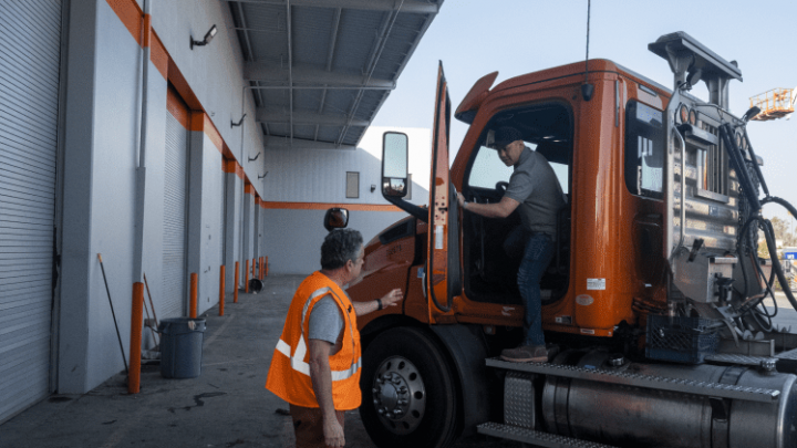 Two fleet-drivers work by an orange semi-truck outside an industrial building, highlighting fleet management on a connected jobsite.