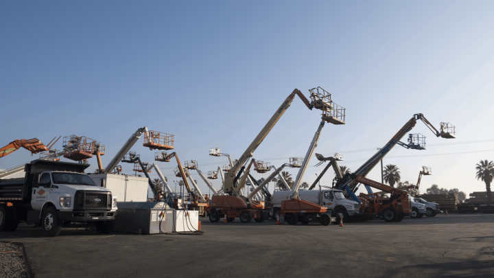 A row of construction vehicles, part of advanced fleet-solutions, with raised boom lifts and cherry pickers parked in a lot under a clear blue sky, with palm trees visible in the background.