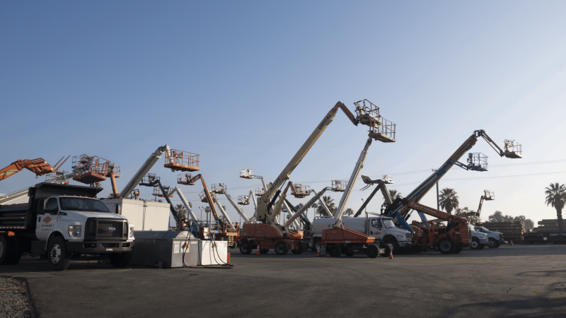 A row of construction vehicles, part of advanced fleet-solutions, with raised boom lifts and cherry pickers parked in a lot under a clear blue sky, with palm trees visible in the background.