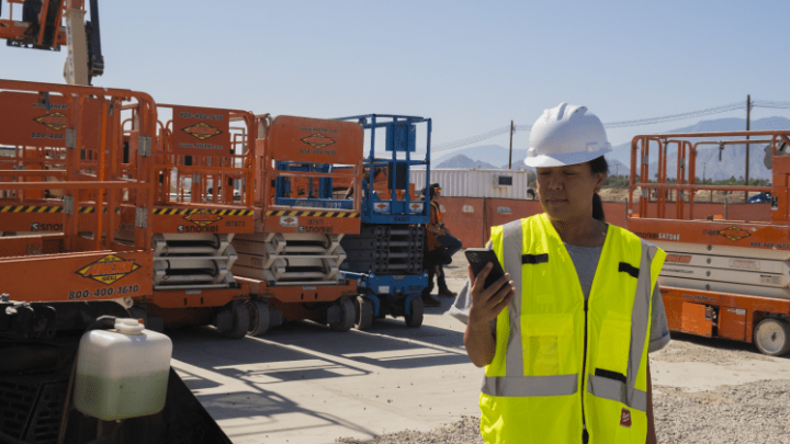 A construction worker in a hard hat and safety vest checks their phone near parked scissor lifts on a sunny day, showing fleet management.