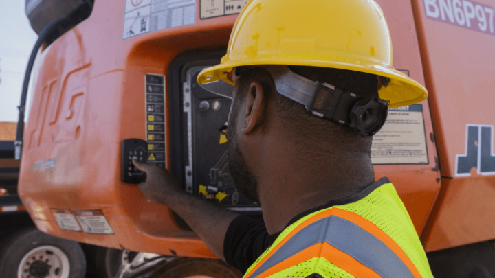 A construction worker in safety gear operates an orange JLG machine at a busy worksite, highlighting construction fleet management.