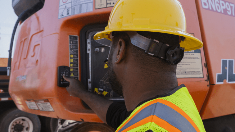 A construction worker in safety gear operates an orange JLG machine at a busy worksite, highlighting construction fleet management.