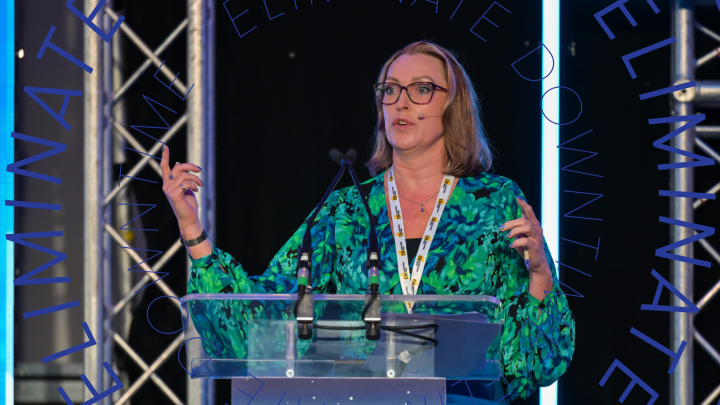 A woman in glasses and a green blouse speaks at a podium about technology, with "ELIMINATE DOWNTIME" signage behind her. Connected jobsite.