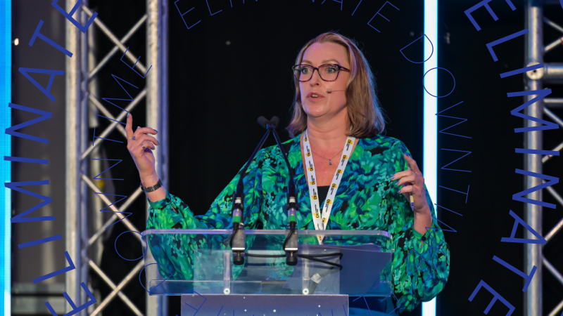 A woman in glasses and a green blouse speaks at a podium about technology, with "ELIMINATE DOWNTIME" signage behind her. Connected jobsite.