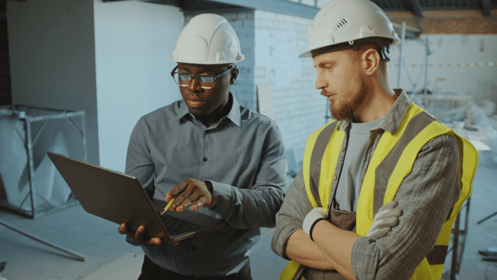Two construction workers in safety helmets review telematics data on a laptop at a construction site surrounded by materials.