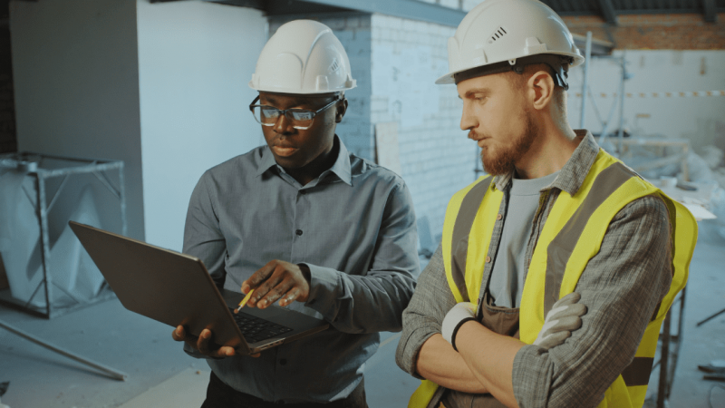 Two construction workers in safety helmets review telematics data on a laptop at a construction site surrounded by materials.