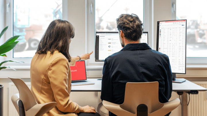Two people at a desk review telematics data on computer monitors, with a potted plant and large windows in the background.