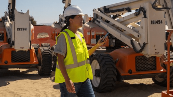Construction worker in hard hat and safety vest walking between heavy machinery at a dusty site, focused on fleet management.