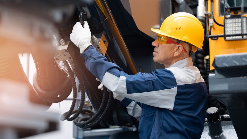 A technician in a yellow hard hat and safety glasses performs maintenance on heavy construction equipment using an OEM component on a connected jobsite.