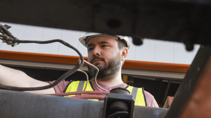 A man in a hard hat and safety vest installs telematics equipment on machinery, low angle with cables in foreground, for connected jobsite.