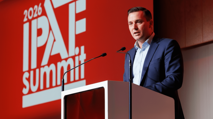 A man in a suit speaks at a podium at the IPAF Summit 2026, backed by a bold red “2026 IPAF summit” display. Fleet management.