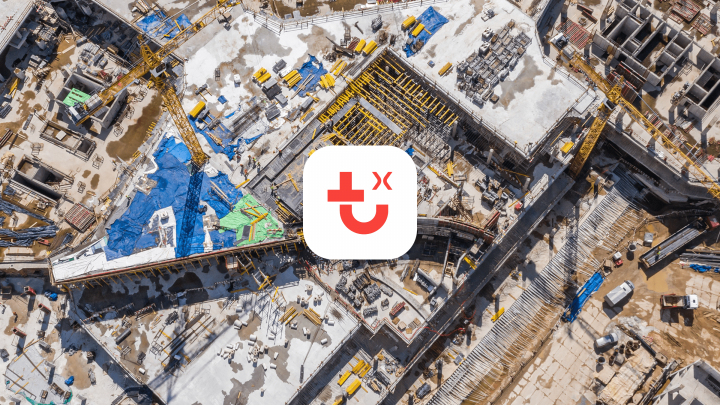 Aerial view of a construction site with slabs, cranes, and machinery. A white app logo with red cross and smiley face is centered.