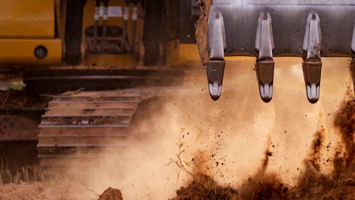 Close-up of an excavator bucket digging into dry soil, dust flying, with yellow body and tracks behind; ideal for construction IoT.