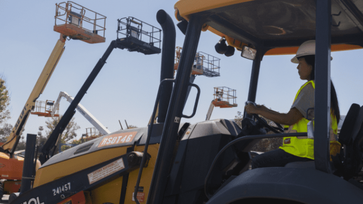 A construction worker in safety gear operates heavy machinery outdoors, with fleet vehicles nearby, highlighting construction equipment tracking.