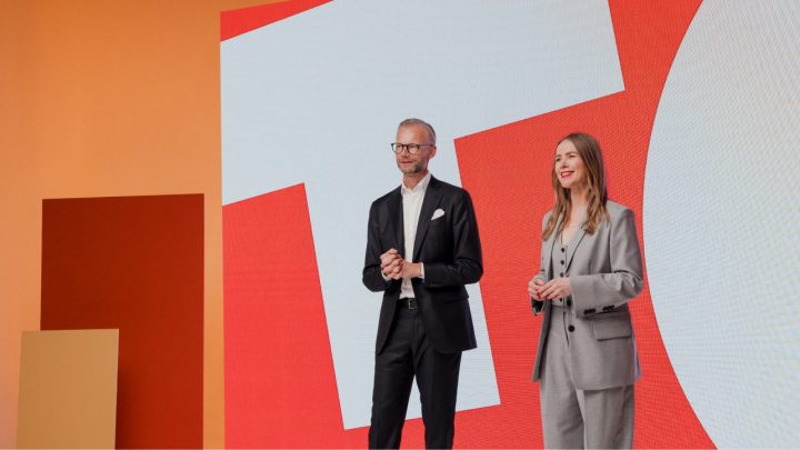 A man in a black suit and a woman in a gray suit smile on stage before a bold geometric backdrop, at a connected jobsite event.