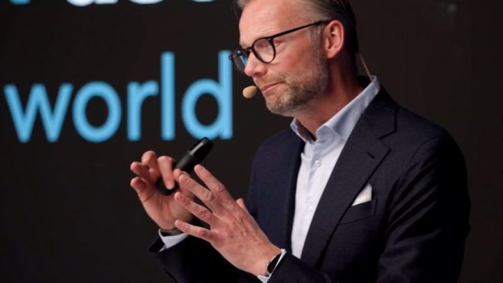 A man in a suit and glasses speaks on stage about safe equipment access, with projected text behind him, related to construction IoT.