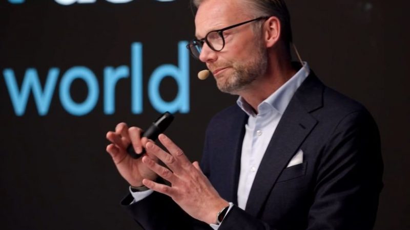 A man in a suit and glasses speaks on stage about safe equipment access, with projected text behind him, related to construction IoT.