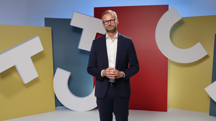 A man in a dark suit and glasses smiles in front of colorful abstract panels and large white letters on a connected jobsite studio set.