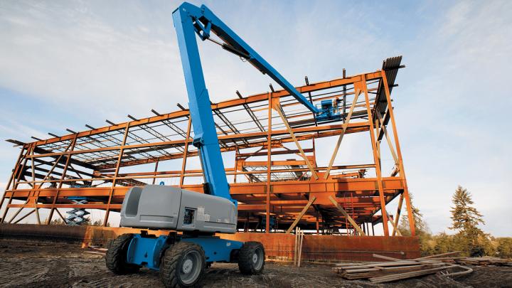 A blue boom lift works by a steel building frame at a connected jobsite, with scattered materials and trees in the background.