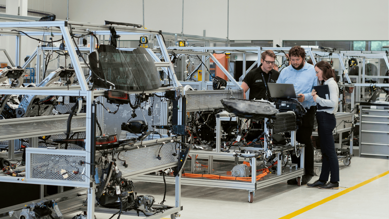 Three people review information on a laptop by a car assembly line in a modern factory, discussing their OEM data platform strategy, illustrating fleet management.