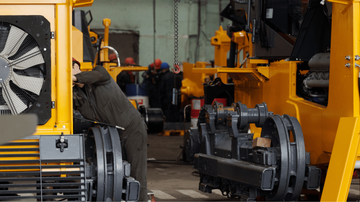 A worker inspects a large yellow machine with construction equipment telematics, surrounded by heavy machinery in a busy factory.