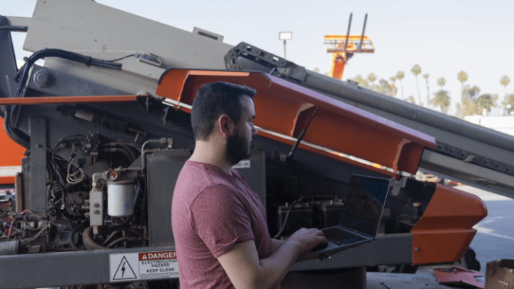 A man outdoors by industrial machinery uses a laptop, monitoring fleet management with palm trees and construction equipment behind him.