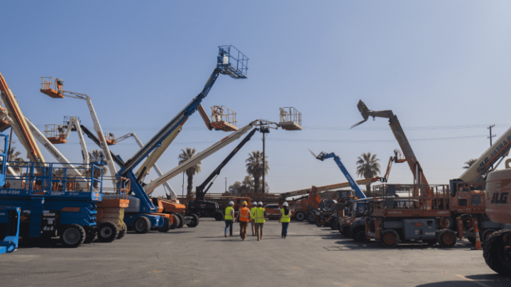 Four people in safety vests walk through a lot of boom and scissor lifts under a blue sky, highlighting construction equipment tracking.