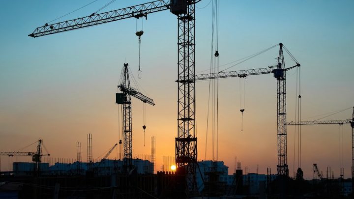 Silhouetted cranes at sunset with the sun behind buildings, capturing growth and construction equipment tracking.