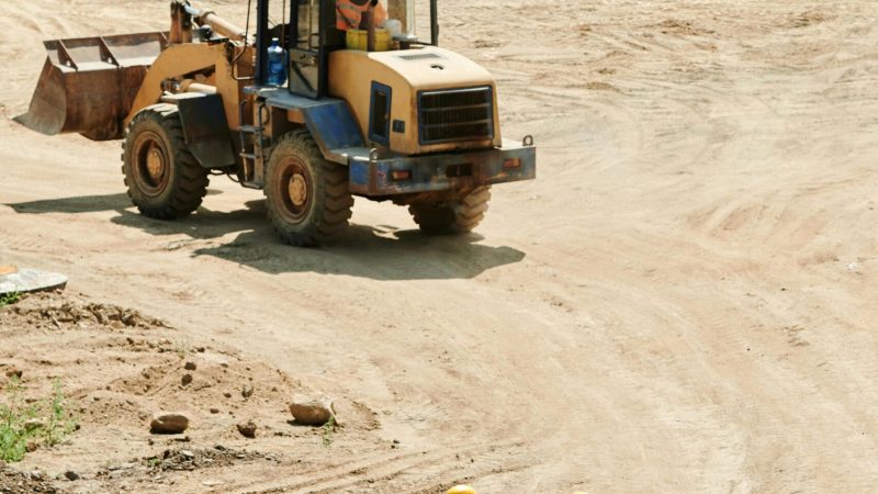 Two workers in yellow vests walk on a dirt path as a front loader operates nearby, highlighting a connected jobsite.